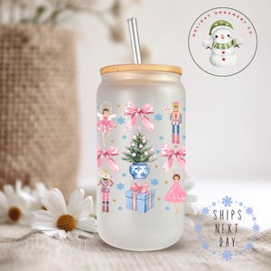 Könnte beinhalten: Ein gefrosteter Glasbecher mit Bambusdeckel und Strohhalm. Der Becher zeigt ein Weihnachtsmotiv mit rosa Schleifen, einem Nussknacker, einer Ballerina, einem Weihnachtsbaum und einer Maus. Der Becher steht auf einer weißen Oberfläche mit weißen Blumen.