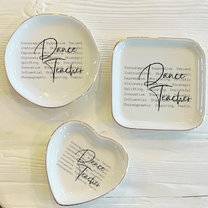 May include: Set of three white ceramic trinket dishes in different shapes: round, square, and heart. Each dish has a gold rim and features the word "Dance Teacher" with supportive adjectives.