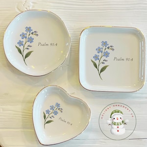 Puede incluir: Tres platos de cerámica blanca con bordes dorados, que presentan una ilustración de flores nomeolvides azules y el texto Psalm 91:4. El conjunto incluye un plato redondo, uno cuadrado y uno en forma de corazón, todos con acabado brillante.