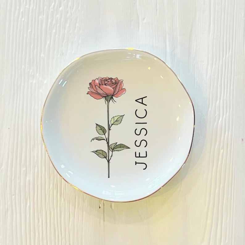 May include: White ceramic trinket dish with a gold rim. The dish features a pink rose with green leaves and the name "JESSICA" printed in black.