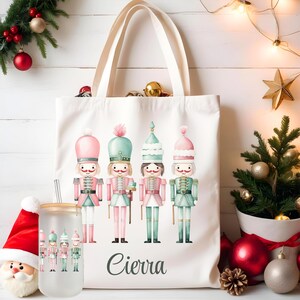 May include: A white canvas tote bag with a Christmas theme featuring five nutcracker figures in pink, green, and blue. The bag has a personalized name, "Cierra", printed on the bottom.