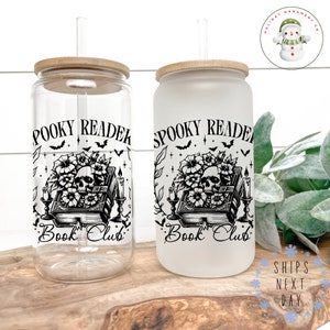 Puede incluir: Dos vasos de vidrio transparentes con tapas de bamb&uacute; y pajitas. Los vasos tienen un gr&aacute;fico en blanco y negro de una calavera, un libro y flores con el texto "Spooky Reader Book Club".