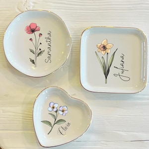May include: Three ceramic trinket dishes with gold trim. The dishes are shaped like a heart, a square, and a circle. Each dish features a different floral design and a name: Olivia, Juliana, and Samantha.