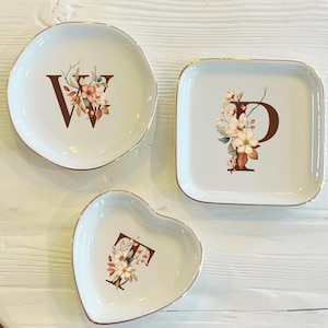 Könnte beinhalten: Drei dekorative weiße Keramikschalen mit goldenen Rändern. Eine runde Schale mit einem "W", eine quadratische Schale mit einem "P" und eine herzförmige Schale mit einem "F". Jede Schale hat ein Blumendesign mit braunen und pfirsichfarbenen Blumen.