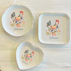 May include: Three decorative ceramic dishes in different shapes: round, square, and heart-shaped. Each dish features a colorful illustration of a rooster and the name "Juliana" in script. The dishes have gold rims.
