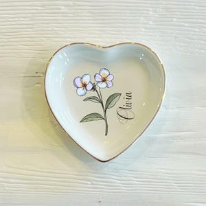 May include: White heart-shaped trinket dish with a gold rim. The dish features a floral design with two light purple flowers and green leaves. The name "Olivia" is written in cursive on the dish.