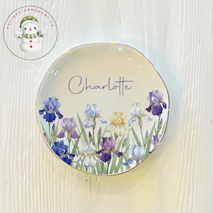 May include: A cream-colored ceramic plate with a gold rim, featuring a floral iris design in shades of blue, purple, and yellow. The name "Charlotte" is written in cursive. The plate is a decorative item.