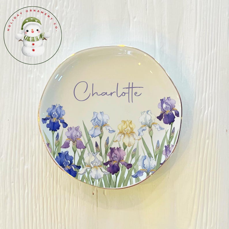 May include: A cream-colored ceramic plate with a gold rim, featuring a floral iris design in shades of blue, purple, and yellow. The name "Charlotte" is written in cursive. The plate is a decorative item.