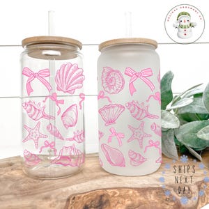 May include: Two glass tumblers with bamboo lids and straws. One is clear, the other frosted. Both are decorated with pink seashell and bow illustrations. The tumblers sit on a wooden board, suggesting a beach or summer theme.