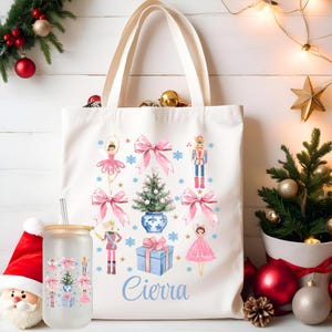 Könnte beinhalten: Eine weiße Canvas-Tragetasche mit einem rosa-blauen Weihnachtsdesign mit einer Ballerina, einem Nussknacker, einem Weihnachtsbaum und einer Geschenkbox. Die Tasche trägt den Namen "Cierra" auf der Vorderseite.