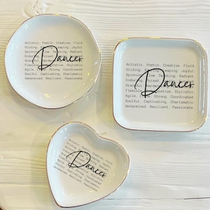 May include: Set of three white ceramic trinket dishes with gold rims. Each dish has the word "Dancer" in a script font, along with descriptive words like "Creative" and "Joyful". The dishes are in different shapes: a circle, a square, and a heart.