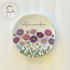 May include: A decorative plate with a floral design featuring purple, pink, and yellow flowers. The plate has a gold rim and the name "Alexandra" is written across the top. The plate is a unique home decor item.