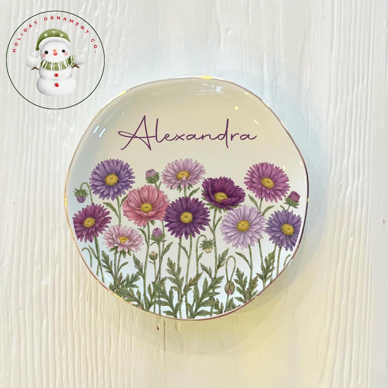 May include: A decorative plate with a floral design featuring purple, pink, and yellow flowers. The plate has a gold rim and the name "Alexandra" is written across the top. The plate is a unique home decor item.