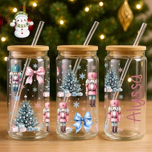 May include: Clear glass tumbler with a wooden lid and a glass straw. The tumbler is decorated with a pink nutcracker, Christmas tree, snowflakes, and a pink bow. One tumbler has the name "Alyssa" in pink script.