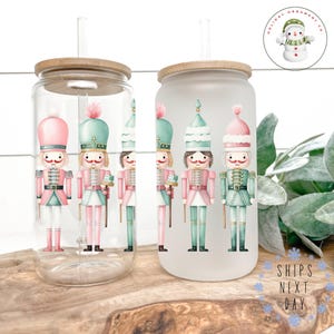Könnte beinhalten: Zwei klare Glasbecher mit einem mattierten Glasbecher. Alle drei Becher haben einen Bambusdeckel und einen wiederverwendbaren Strohhalm. Die Becher sind mit einem Aquarelldesign von vier Nussknackern in Rosa, Grün und Weiß verziert.
