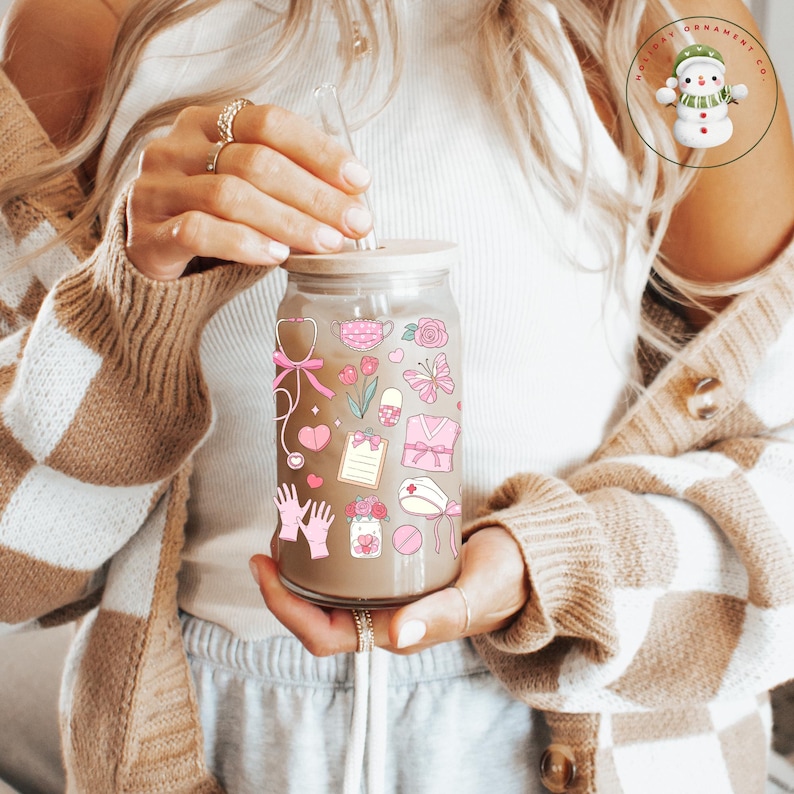 May include: A clear glass tumbler with a wooden lid and a straw, decorated with pink medical-themed stickers. The stickers include a stethoscope, scrubs, and a nurse's hat. The tumbler is held by a person.