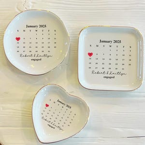 May include: Three white ceramic ring dishes with gold trim. Each dish features a calendar for January 2025 with a red heart marking the 12th. The text "Robert & Kaitlyn engaged" is printed below the calendar.