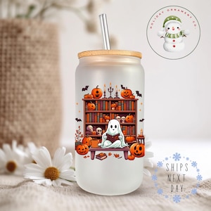 Op de afbeelding: Een matglazen tumbler met een Halloween-thema. Het ontwerp toont een geest die een boek leest voor een boekenkast vol boeken en pompoenen. De tumbler heeft een houten deksel en een rietje.