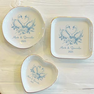 Könnte beinhalten: Drei weiße Keramik-Ringschalen mit goldenen Rändern. Jede Schale zeigt eine blaue Illustration von zwei Schwänen, die eine Herzform bilden, mit floralen Akzenten. Der Text "Mark & Danielle 2025" ist auf jeder Schale aufgedruckt.