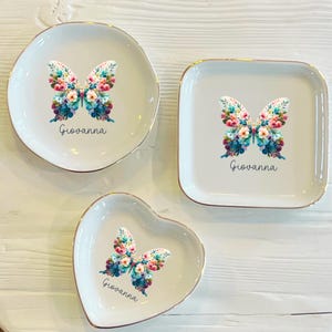 Puede incluir: Tres platos de cerámica para anillos con un borde dorado. Cada plato presenta una mariposa colorida con detalles florales y el nombre "Giovanna" impreso debajo. Los platos tienen forma de círculo, cuadrado y corazón.