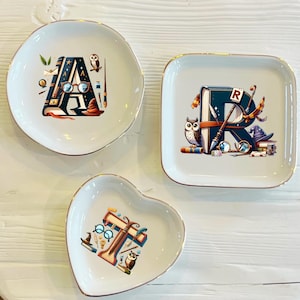 Puede incluir: Tres platos de cerámica para joyas con ribete dorado, cada uno con una letra diferente del alfabeto, A, R y T, decorados con una ilustración caprichosa de un libro, un búho y otros objetos mágicos.