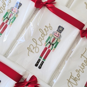 May include: Set of white kitchen towels with a red ribbon and bow. Each towel features an embroidered nutcracker design and a personalized name in gold thread. The names on the towels are "Blades" and "Christmas".