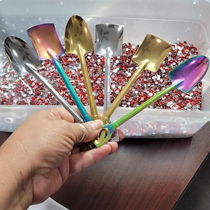 May include: Six miniature shovels with different colored handles and metallic finishes. The shovels are arranged in a fan shape and are sitting in a clear plastic container filled with red and silver rhinestones. The container is on a wooden surface.