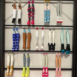 Può includere: Una collezione di orecchini a forma di gambe di bambola esposti su una cornice nera. Gli orecchini presentano vari stili, tra cui calzini bianchi con scarpe nere, gambe rosa avvolte da serpenti, sandali gladiatore blu e stivali marroni.