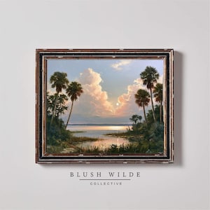 May include: A framed landscape painting depicting a sunset over a calm body of water, framed by tall palm trees and lush vegetation. The sky features large, soft clouds illuminated by a golden sun. The text BLUSH WILDE COLLECTIVE appears below.