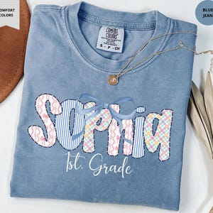 May include: Blue jean Comfort Colors t-shirt with the name "Sophia" in a patchwork design, a blue bow, and the text "1st. Grade". A gold necklace with a pendant is on the shirt.
