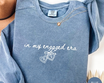 May include: A blue jean colored crewneck sweatshirt featuring the white text in my engaged era above a graphic of two connected hearts with the letters M and B inside. The garment has a ribbed collar and long sleeves with a Comfort Colors label.