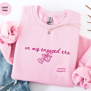 Light Pink Comfy Sweatshirt with custom raspberry text "in my engaged era" and heart lock graphic, perfect for brides-to-be and engagement gifts.