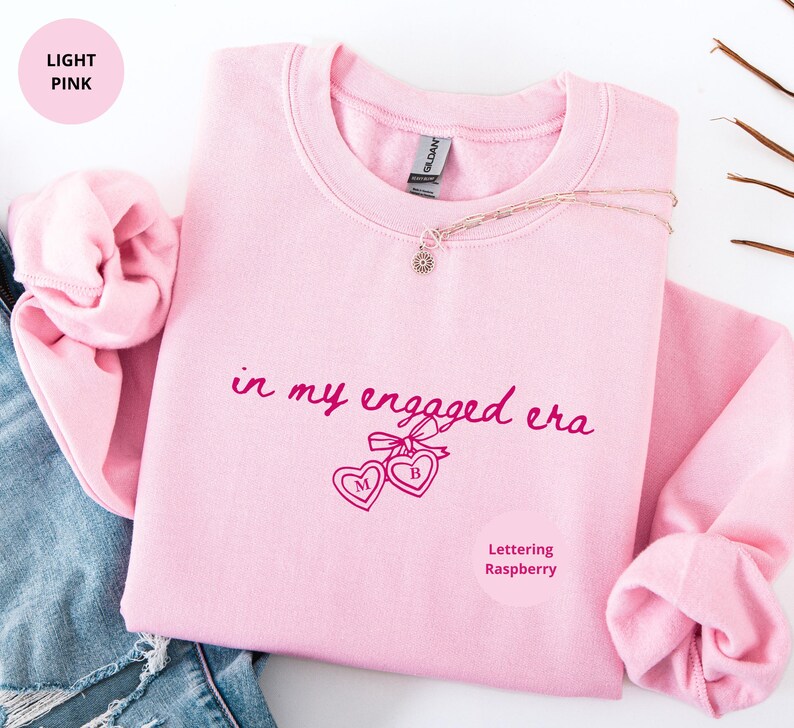 Light Pink Comfy Sweatshirt with custom raspberry text "in my engaged era" and heart lock graphic, perfect for brides-to-be and engagement gifts.