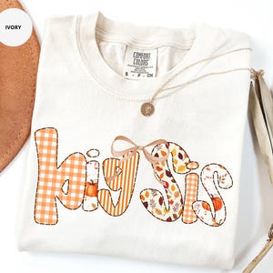 May include: Ivory-colored t-shirt with the words "Hey Sis" in a patchwork design. The letters feature orange gingham, stripes, and fall leaf patterns. A small bow is between the words. A gold necklace with a pendant is on the shirt.