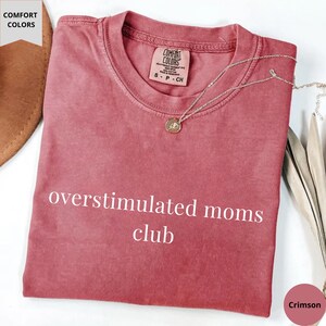 May include: Crimson-colored t-shirt with the text "overstimulated moms club." The shirt has a crew neck and short sleeves. A silver necklace with a small pendant rests on the shirt. The shirt is made of a soft material.