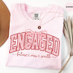 May include: A light pink t-shirt featuring the word ENGAGED in large, red-outlined block letters with a patterned fill of small hearts and bows. A light pink bow graphic sits above the text, and the words future mrs smith appear below.