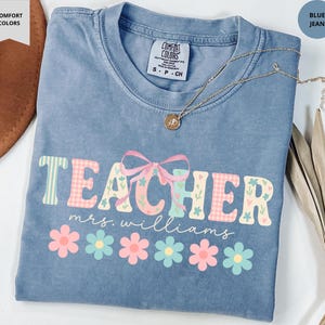 May include: Blue jean-colored t-shirt with the word "TEACHER" in a floral and gingham design, with "mrs. williams" below. A gold necklace with a pendant and a pink bow are also visible. The shirt is a Comfort Colors brand.