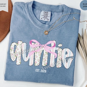 May include: Blue jean-colored t-shirt with the word "Auntie" in a floral and striped design, accented by a pink bow. The text "EST. 2026" is below the word. A gold necklace with a pendant is on the shirt.