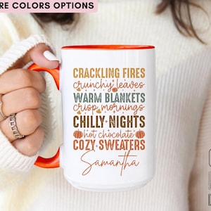 May include: White ceramic mug with an orange interior and handle. The mug features text in various colors, including "CRACKLING FIRES," "WARM BLANKETS," and "COZY SWEATERS." The name "Samantha" is also printed on the mug.
