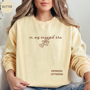 Butter Comfort Colors® sweatshirt with custom espresso text "in my engaged era" and heart lock graphic, perfect for brides-to-be and engagement gifts.