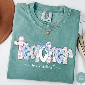 May include: Light green Comfort Colors t-shirt with the word "Teacher" in a patchwork design featuring bows and stripes. The name "mrs. michael" is written below the word "Teacher". A silver necklace with a pendant is on the shirt.