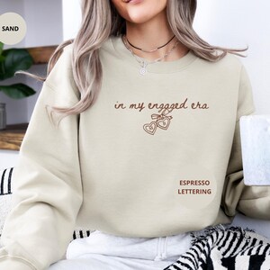 Sand Sweater with custom espresso text "in my engaged era" and heart lock graphic, perfect for brides-to-be and engagement gifts.