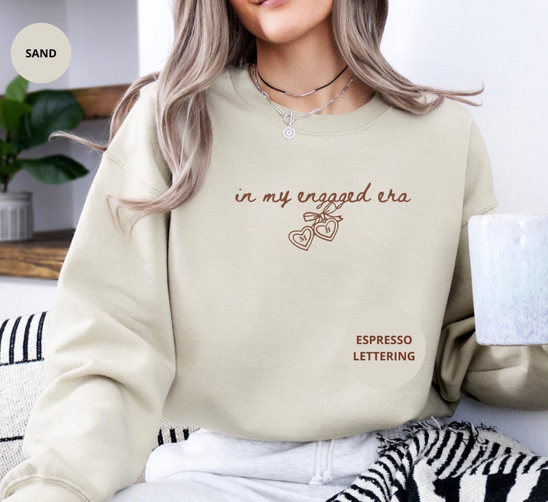 Sand Sweater with custom espresso text "in my engaged era" and heart lock graphic, perfect for brides-to-be and engagement gifts.