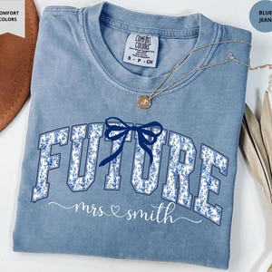 May include: A blue jean Comfort Colors t-shirt with the word "FUTURE" in a floral design, a blue bow, and the words "mrs & smith" in script. A gold necklace with a pendant is on the shirt.