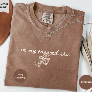 Espresso Comfort Colors® t-shirt with custom ivory text "in my engaged era" and heart lock graphic, perfect for brides-to-be and engagement gifts.