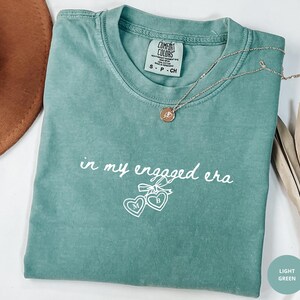 Light Green Comfort Colors® t-shirt with custom white text "in my engaged era" and heart lock graphic, perfect for brides-to-be and engagement gifts.