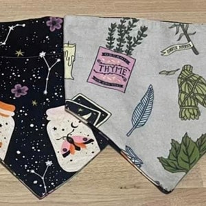 May include: Two fabric bandanas with different designs. One bandana is black with white stars, jars, and a butterfly. The other bandana is light gray with a pink and white "THYME" label, a green leaf, and a white feather. Both bandanas have a triangular shape.