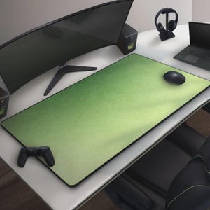 May include: A large, rectangular green mouse pad with a black border sits on a white desk. A black computer mouse rests on the pad. A black game controller is on the desk. A curved monitor, speakers, and a laptop are also visible.
