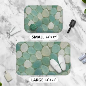 May include: Two bath mats with a pebble design in shades of green and white. The smaller mat is 24 inches by 17 inches and the larger mat is 34 inches by 21 inches.