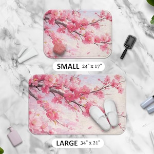 Puede incluir: Dos alfombras de baño florales de color rosa y blanco con las palabras "SMALL 24" x 17" y "LARGE 34" x 21" impresas en ellas. Las alfombras presentan un diseño de estilo acuarela de flores de cerezo rosas sobre un fondo blanco.